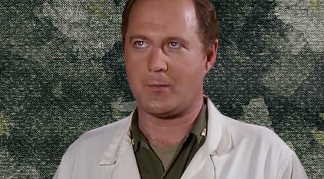 David Ogden Stiers in his army uniform and lab coat against a camoflauge background from MASH